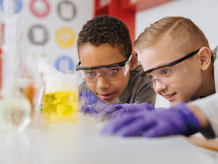 Teenage schoolboys in safety goggles leaning closely to the table and watching chemical reaction going on in the flask