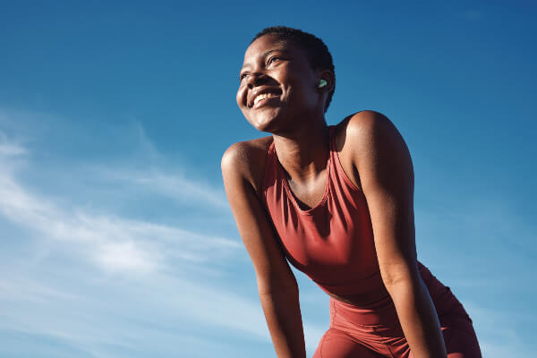 Fitness, black woman and happy athlete smile after running, exercise and marathon training workout. Blue sky, summer sports and run of a African runner breathing with happiness from sport outdoor.