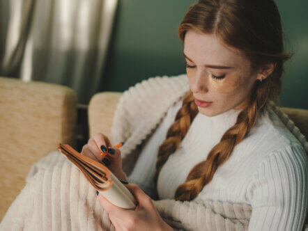 Young minded woman wrapped in plaid writing down memories in notebook sitting on sofa at home spending free spare time in living room.