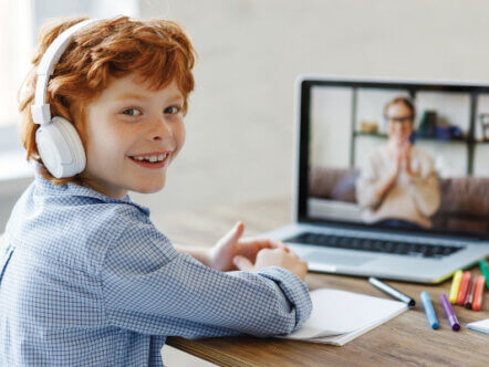 Happy boy in headphones smiling and looking at camera while making video call to teacher during online lesson at home